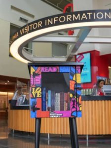 Little Free Library at with colorful painting below the sign for the Visitor Information booth